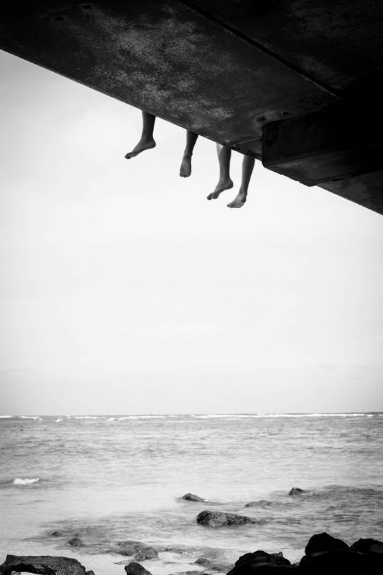 Two pairs of feet dangling off a bridge over the ocean
